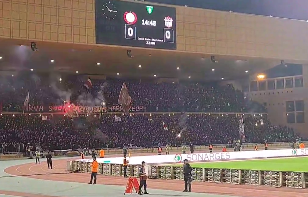 Football fans cheering with flares, smoke, and flags in the stands at the Grand Stade de Marrakech.