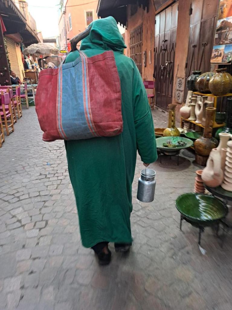 A local person in a green hooded djellaba walking down a cobbled Marrakesh street carrying a striped bag and a metal milk container.v