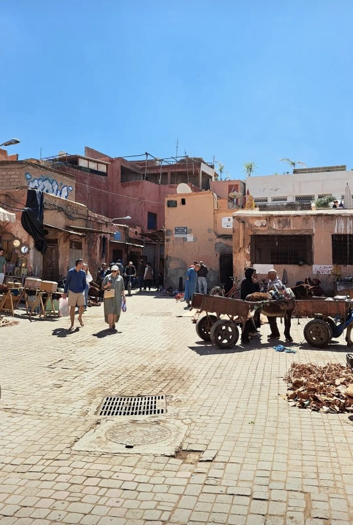 People walking through a sunny square in the Marrakesh Medina with a wooden donkey cart in the foreground.