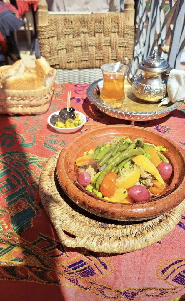 A traditional Moroccan meat and vegetable tagine served on a colorful table with mint tea, bread, and olives.